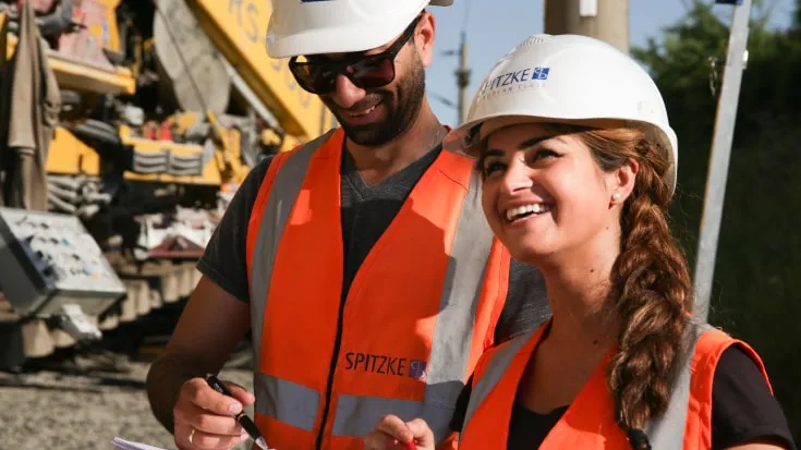 Zwei Bauarbeiter in oranger Schutzkleidung stehen lachend auf einer Baustelle.